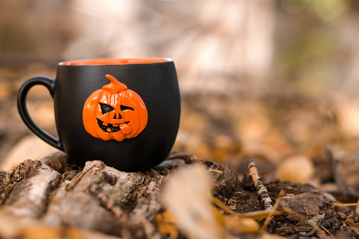 Black mug with a hot tea stands on the ground among the fallen yellow autumn leaves and branches.The mug is decorated with a pumpkin jack o'lantern.Halloween concept.Selective focus, copy space.