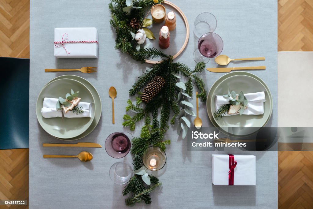 New Year's Eve Preparations: Top View of Elegant Christmas Table Setting for Two People Beautiful holiday table setting with golden cutlery and green porcelain plate on a gray table with Christmas decoration and presents. Party Host Stock Photo