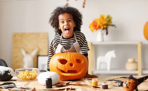 Photo of Playful little afro american boy holding carved orange pumpkin Jack o lantern while standing behind table