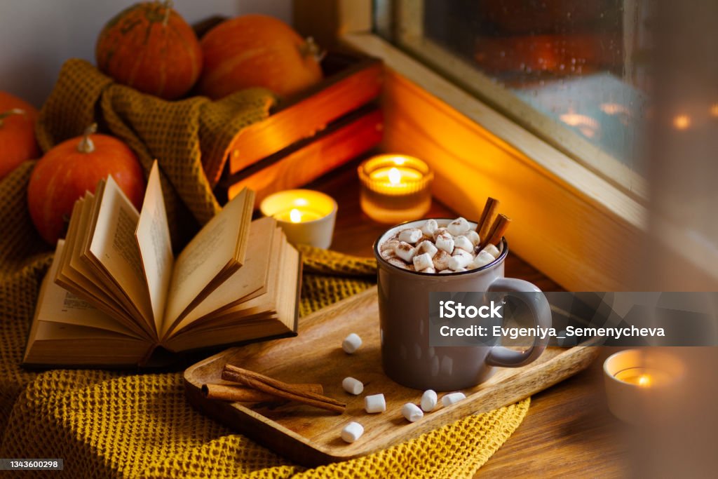 a cup of hot chocolate with marshmallows is on the window Mug of cocoa or hot chocolate with marshmallows next to the window with candles, pumpkins, book and warm blanket. Cozy home atmosphere in rainy autumn day. Нygge lifestyle concept Candle Stock Photo