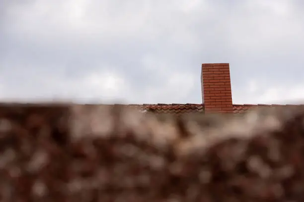 Red brick chimney on a tiled house roof viewed over blurred foliage in the foreground against a cloudy sky