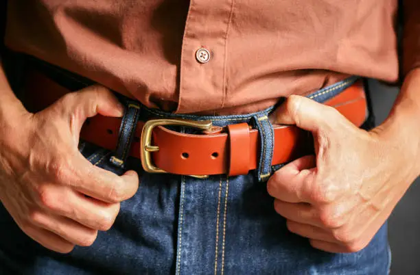 Photo of A man in jeans and a shirt fastens his leather belt.