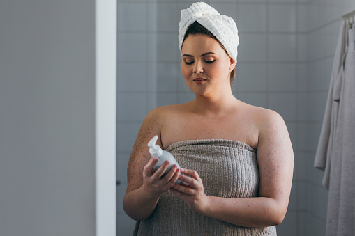 A young overweight woman reading the ingredients of her body lotion after a shower in the morning