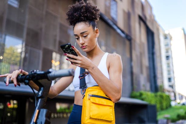 joven de raza mixta de pie en la calle con scooter eléctrico mientras usa un teléfono inteligente - ciclomotor fotografías e imágenes de stock