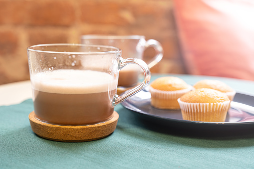 A glass cup of hot cocoa or cappuccino on a cork stand, three muffins on a plate, standing on a table, illuminated by light from a window. Breakfast for two. In a cafe for a morning cup of coffee.