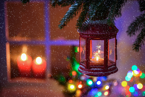 Close up of a red lantern hanging from a fir branch on a snowing night. A window lit by candles and defocused lights are in background. Christmas background. Copy space.