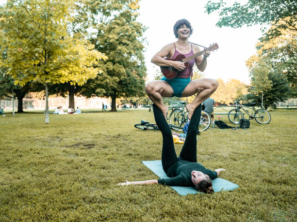 Couple of young women practising Acro Yoga. One woman balancing in the air with guitar while other is on the ground holding her above. Both dressed in casual sport clothing. Various backgrounds and nationalities. Exterior of public park in sunset.