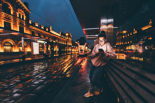 Happy lady with glasses and mobile phone listens to music sitting on city transport stop in illuminated city street with blurred lights at rainy night