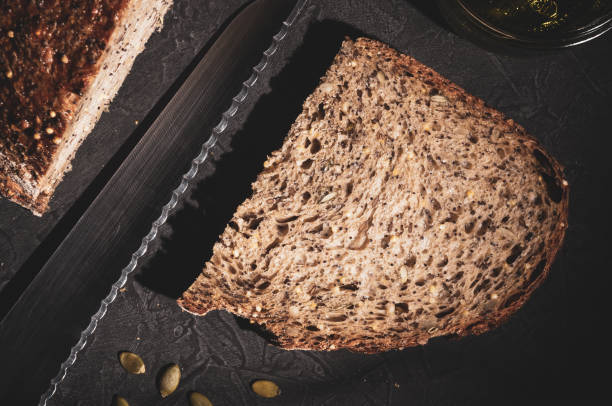 slice of rustic natural yeast-free bread with flax, poppy seeds, sesame seeds, millet, pumpkin and sunflower seeds, with olive oil in a glass jar, and a knife, on a black background, hard light, photo in a low key rebanada de pan rústico natural sin levadura con lino, semillas de amapola, semillas de sésamo, semillas de mijo, calabaza y girasol, con aceite de oliva en un frasco de vidrio, y un cuchillo, sobre un fondo negro, luz dura, foto en clave baja.