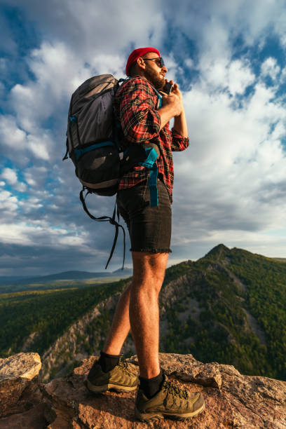 A male traveler in a red hat with a backpack is talking on the phone against the background of mountains. A man in hiking clothes talks on a smartphone at sunset in the mountains. Phone conversation stock photo