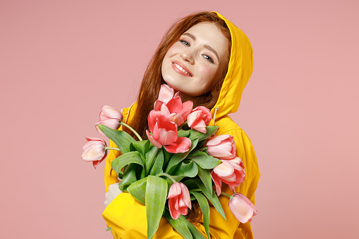 Redhead young satisfied pretty woman in yellow waterproof hood raincoat outerwear giving tulips flower bouquet isolated on pastel pink background studio Lifestyle spring weather blossom season concept
