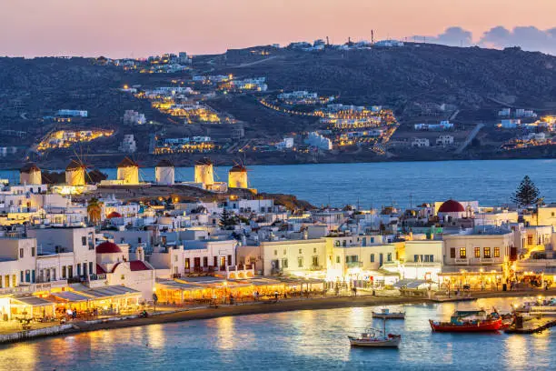 Photo of Chora port of Mykonos island with famous windmills, ships and yachts during colorful sunset. Aegean sea, Greece