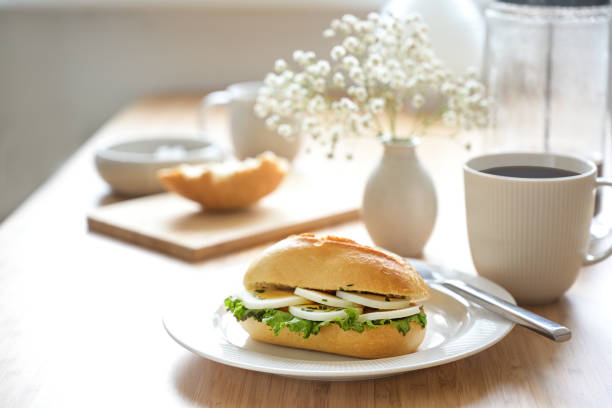 Breakfast table set with egg slices in a sandwich roll, coffee cup and a small flower bouquet, light background with copy space, selected focus, narrow depth of field