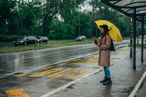 Beautiful woman using a smartphone and holding a yellow umbrella while on the bus stop in the city on a rainy day
