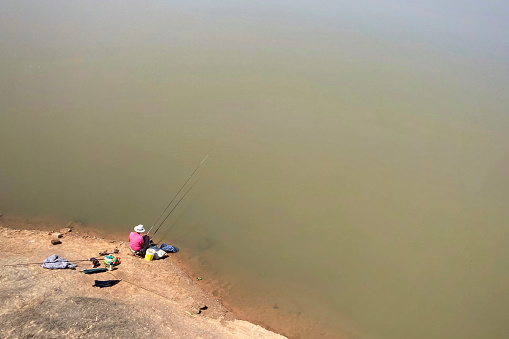 Photograph of an aerial view of a fisherman at a lake. The lake looks calm, in a rural area in Brazil. Behind the fisherman there ar esmole fishing equipments.