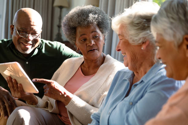 tre felici donne anziane diverse e un amico maschio afroamericano seduto sul divano e usando il tablet - retirement living foto e immagini stock