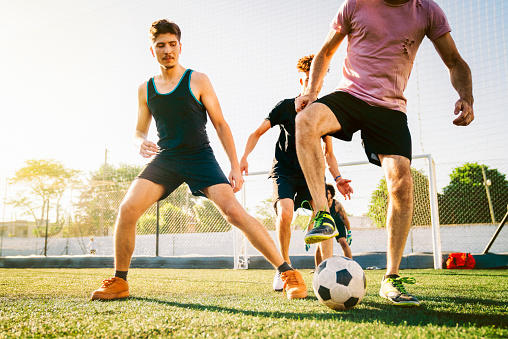Mature man defending ball while playing soccer with young male friends. Athletes are practicing together during sunny day. They are at sports field.