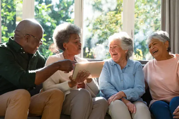 Photo of Three happy diverse senior woman and african american male friend sitting on sofa and using tablet