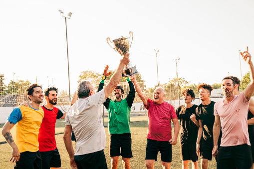 Happy senior man raising trophy while celebrating with male friends. Players are cheering together at field. They are enjoying victory.