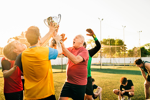 Happy male players holding trophy while celebrating together during sunny day. Athletes are cheering after soccer game. They are at training ground.