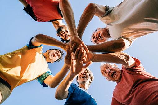 Happy male soccer players stacking hands against sky. Men are huddling during sunny day. They are in sports clothing.