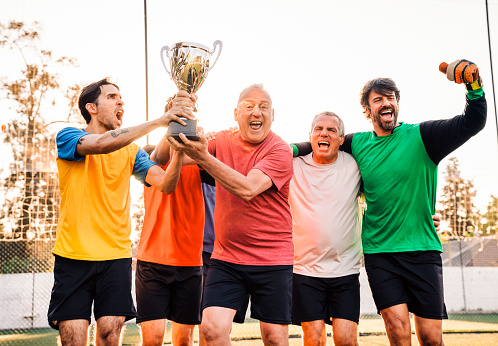 Portrait of happy senior man holding trophy while celebrating with team. Male soccer players are cheering together. They are enjoying at soccer field.