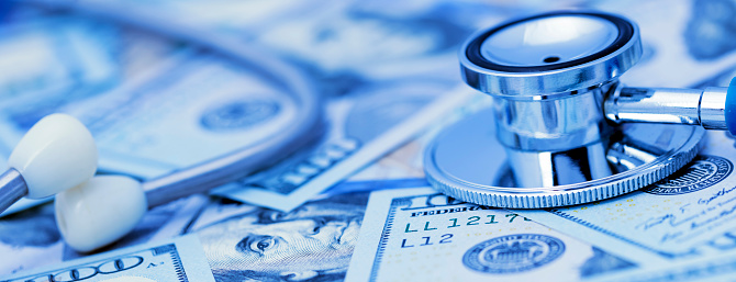 Close-up of a stethoscope on a heap US $100 dollars bills. Blue toned image.