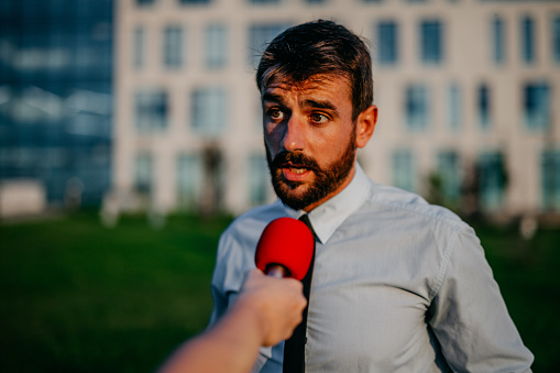 Spontaneous portrait of a handsome bearded young adult lawyer, an unrecognizable journalist holding a red microphone, while a guy is talking outdoors, on a beautiful sunny day. He’s wearing a shirt and a tie.