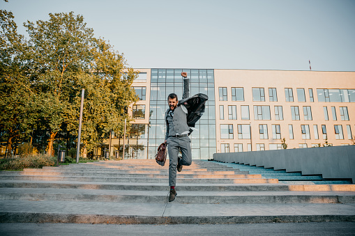 Embracing and strong photo of a handsome bearded young adult businessman, a good lawyer, jumping down the stairs, in front of the court, celebrating his won case while carrying a briefcase in a stylish gray suit. Looking sharp and happy like a kid at the same time