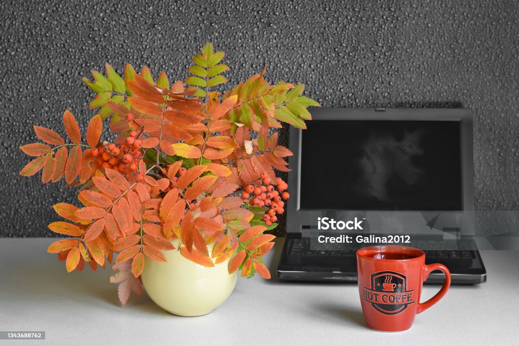 A mug of hot coffee with steam, on the background of a computer and a bouquet of autumn leaves A mug of hot coffee with steam, against the background of a computer and a bouquet of autumn leaves in a vase. Selective focus Autumn Leaf Color Stock Photo