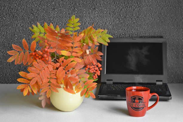 A mug of hot coffee with steam, on the background of a computer and a bouquet of autumn leaves stock photo