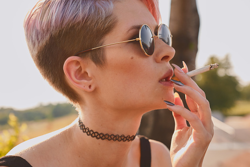 Portrait of daring young woman with short pink hair in round glasses outdoors, she is smoking a cigarette, holding it in hands, inhaling smoke