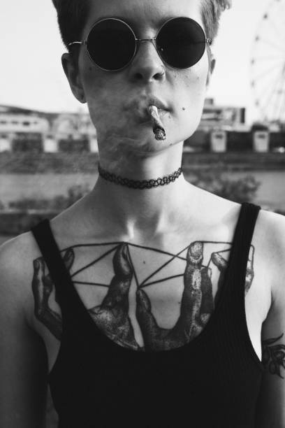 Daring young woman smoking cigarette outdoors Portrait of smoking woman. She is holding cigarette in mouth handsfree, she has short hair, wearing round glasses, tattooes on her chest rock musician photos stock pictures, royalty-free photos & images