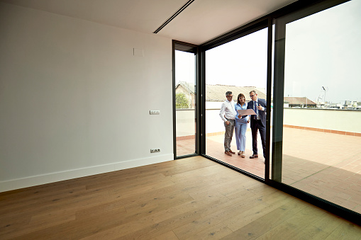 View from apartment interior through open sliding door to male Real Estate Agent and mature couple standing outdoors on deck looking at property details.