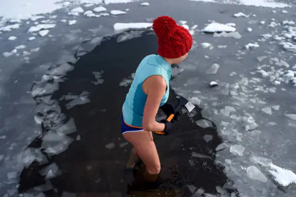 Photo of High angle view of active senior woman in swimsuit breaking ice with axe outdoors in winter, cold therapy concept.