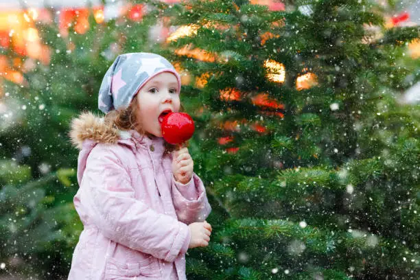 Cute little smiling preschool girl on German Christmas market. Happy child in winter clothes eating sweet sugared glazed xmas apple on with lights on background. Family, tradition, celebration concept.
