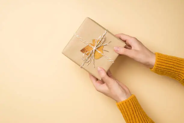 Photo of First person top view photo of hands in yellow pullover giving craft paper giftbox with twine bow and yellow autumn leaf on isolated beige background with copyspace