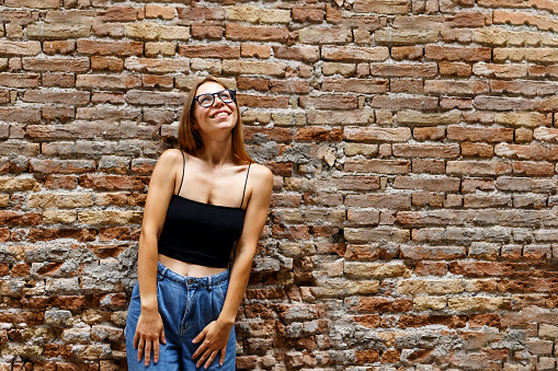 A hipster girl with long brown hair wearing a denim is looking aside while standing on a brick wall background on a street. Empty space for text o design.