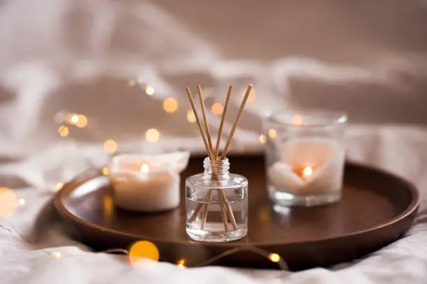 Home perfume in glass bottle with wood sticks, scented burn candles  tray in bedroom close up. Aromatherapy cozy atmosphere lifestyle. Winter warm xmas season.