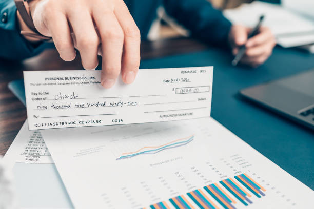 la mano di un uomo d'affari che dà assegno al cliente e banconota da un dollaro, moneta, laptop e grafico sulla scrivania dell'ufficio. pagamento tramite assegno, busta paga, concetto di busta paga. - order pad foto e immagini stock