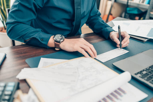 geschäftsmann handschrift und signieren weißer blanker bankscheckbuch und dollarschein, münze, laptop und diagramm auf dem schreibtisch im büro. zahlung per scheck, gehaltsscheck, gehaltsabrechnungskonzept. - scheck stock-fotos und bilder