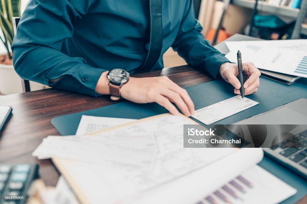 Business man hand writing and signing white blank bank check book and dollar bill, coin, laptop and graph chart on the desk at office. Payment by check, paycheck, payroll concept. Check - Financial Item Stock Photo