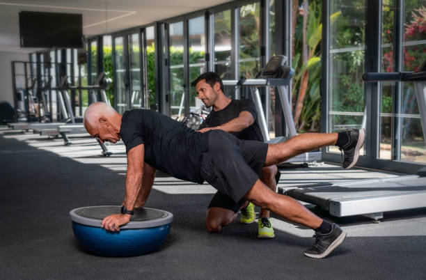 un anziano uomo caucasico, in palestra, con un personal trainer fitness, in riabilitazione, facendo esercizi per l'equilibrio su una palla ginnica. - fitness instructor foto e immagini stock