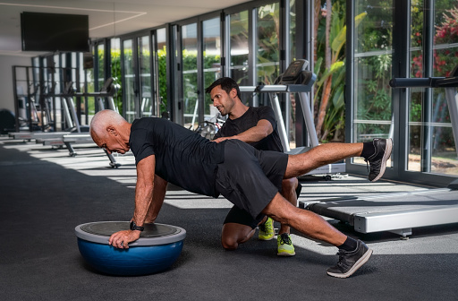 An elderly Caucasian man, in the gym, with a personal fitness trainer, in rehabilitation, doing exercises for balance on a gymnastic ball. High quality photo