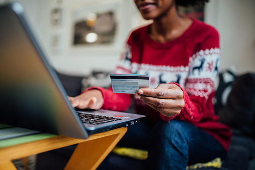Close up of an unrecognizable African American woman enjoying herself at home during winter holidays, using a laptop to shop online and holding a credit card in her hand