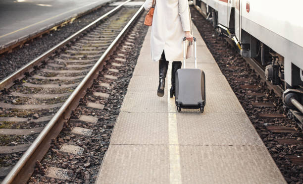istock Young woman in white coat pulling her black trolley luggage behind on train platform, view from behind only anonymous lower part of body visible 1343793180