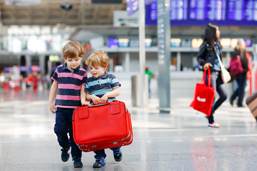Two little sibling boys having fun and going on vacations trip with suitcase at airport, indoors. Happy twins brothers walking to check-in or boarding. Family on flight, journey on plane.