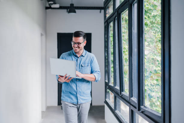 giovane allegro allegro bello gioioso manager imprenditore imprenditore piccolo imprenditore impiegato lavoratore con gli occhiali sorridente mentre lavora con il suo laptop camminando nel corridoio del corridoio dell'ufficio - moving up looking men people foto e immagini stock