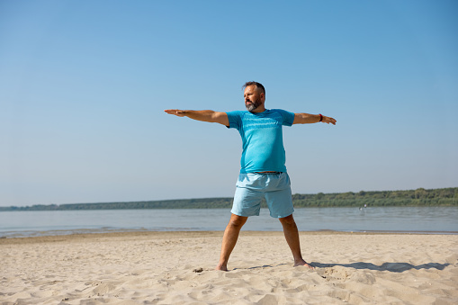 Mid adult man in blue shorts and T-shirt doing yoga exercise on sandy beach, arms outstretched and legs spread