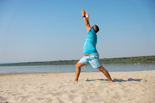 Mid adult man in blue shorts and T-shirt doing yoga exercise on sandy beach, arms raised, looking up, lunge posture, element of Sun Salutation